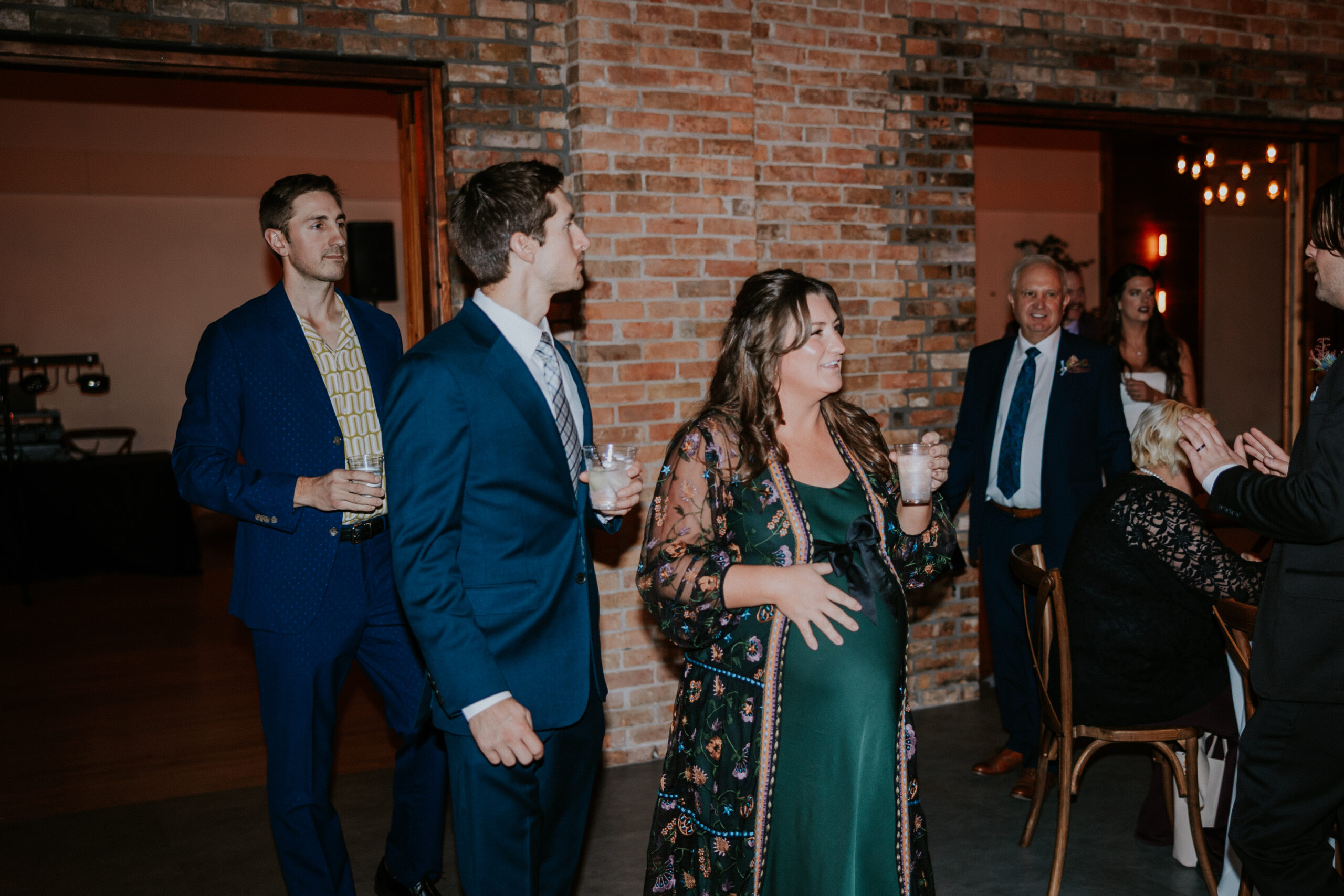 A guest in a green dress with a floral shawl stands beside loved ones near a rustic brick wall at a wedding in Minnesota.