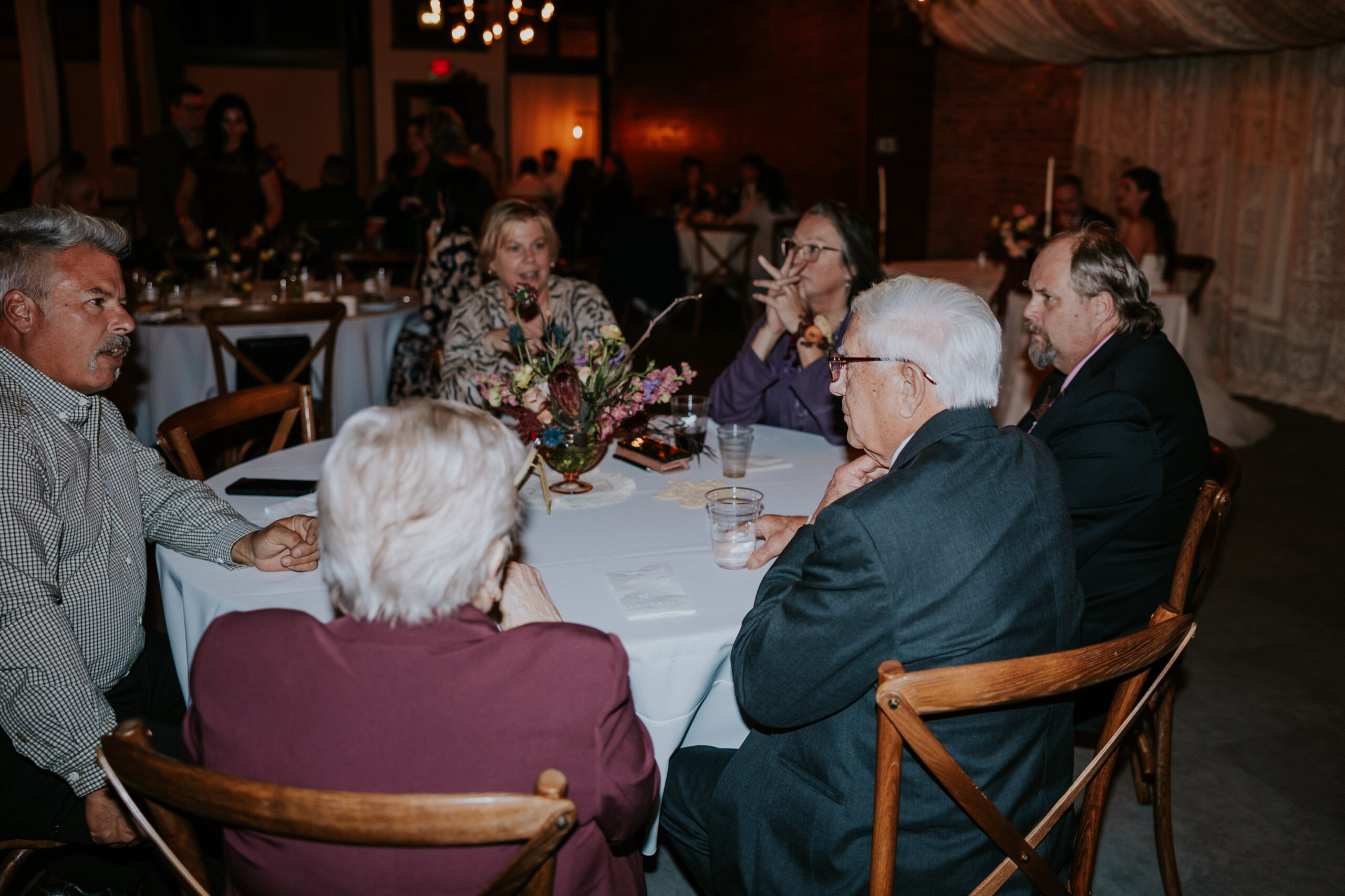 Guests enjoying conversation around a candlelit table decorated with flowers during a cozy indoor wedding celebration.