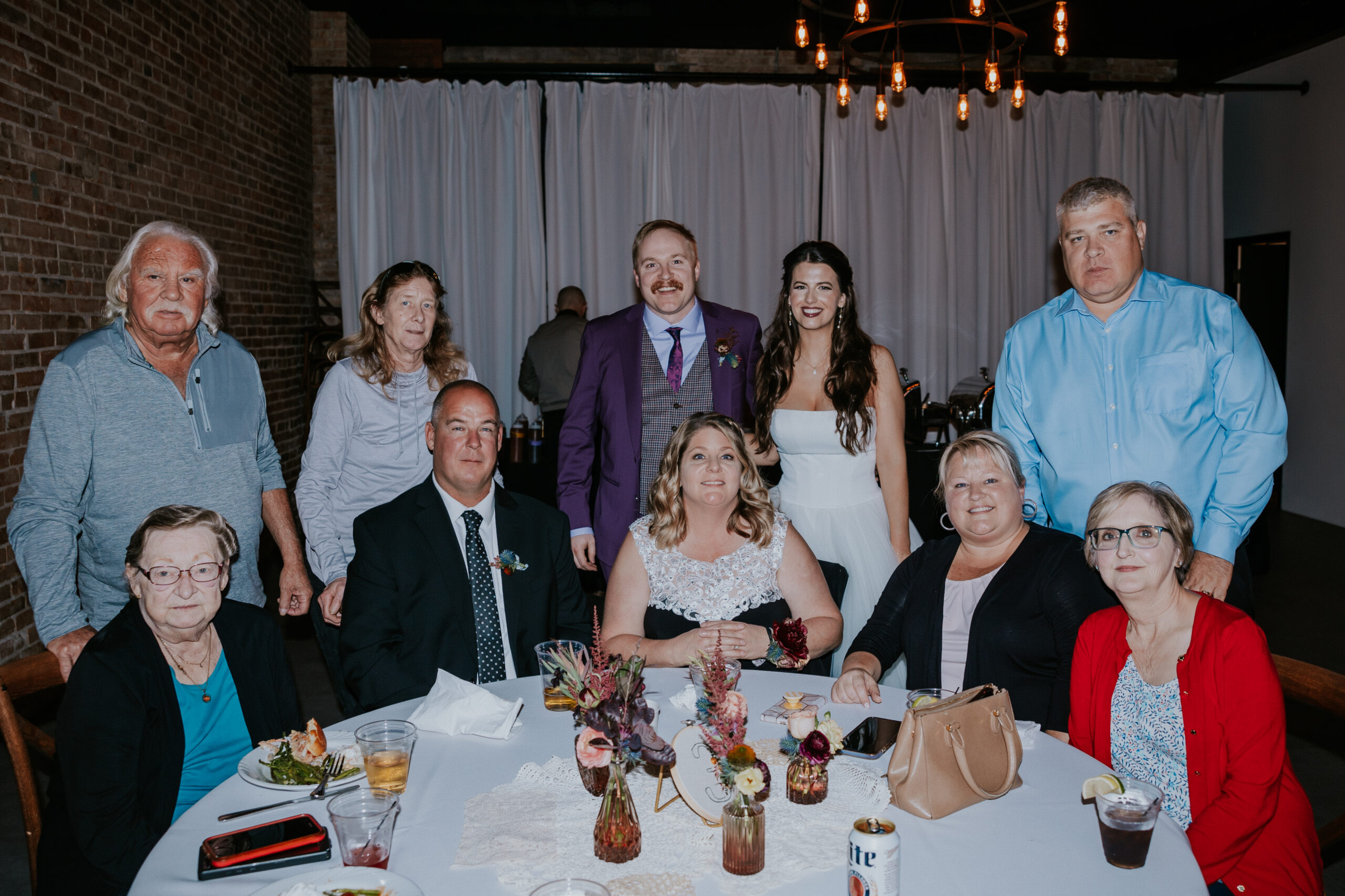The bride and groom pose with family members during their wedding in Minnesota, surrounded by love and warm smiles inside a cozy indoor venue.