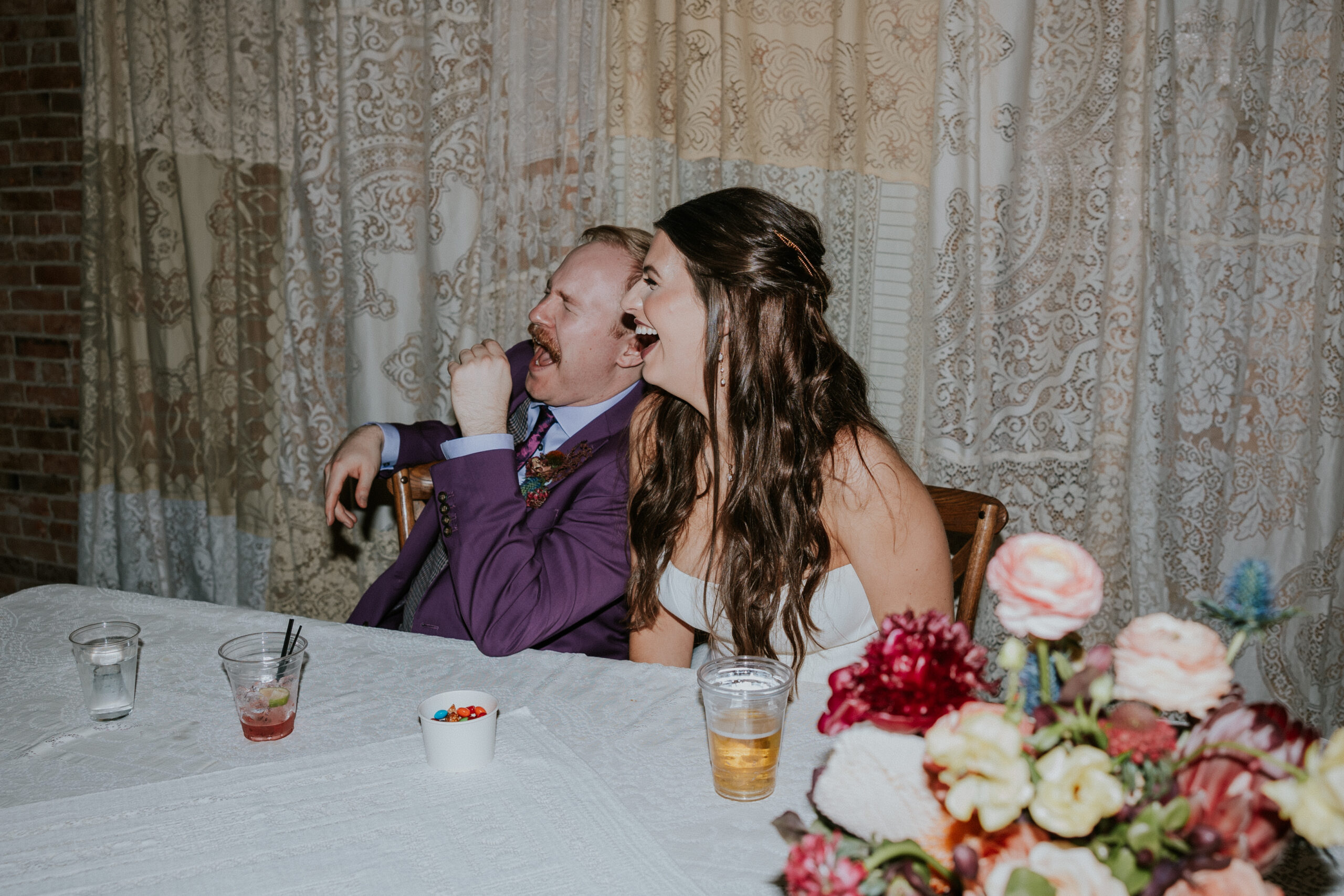 The bride and groom laugh together while seated at their sweetheart table, enjoying heartfelt speeches at their wedding in Minnesota.