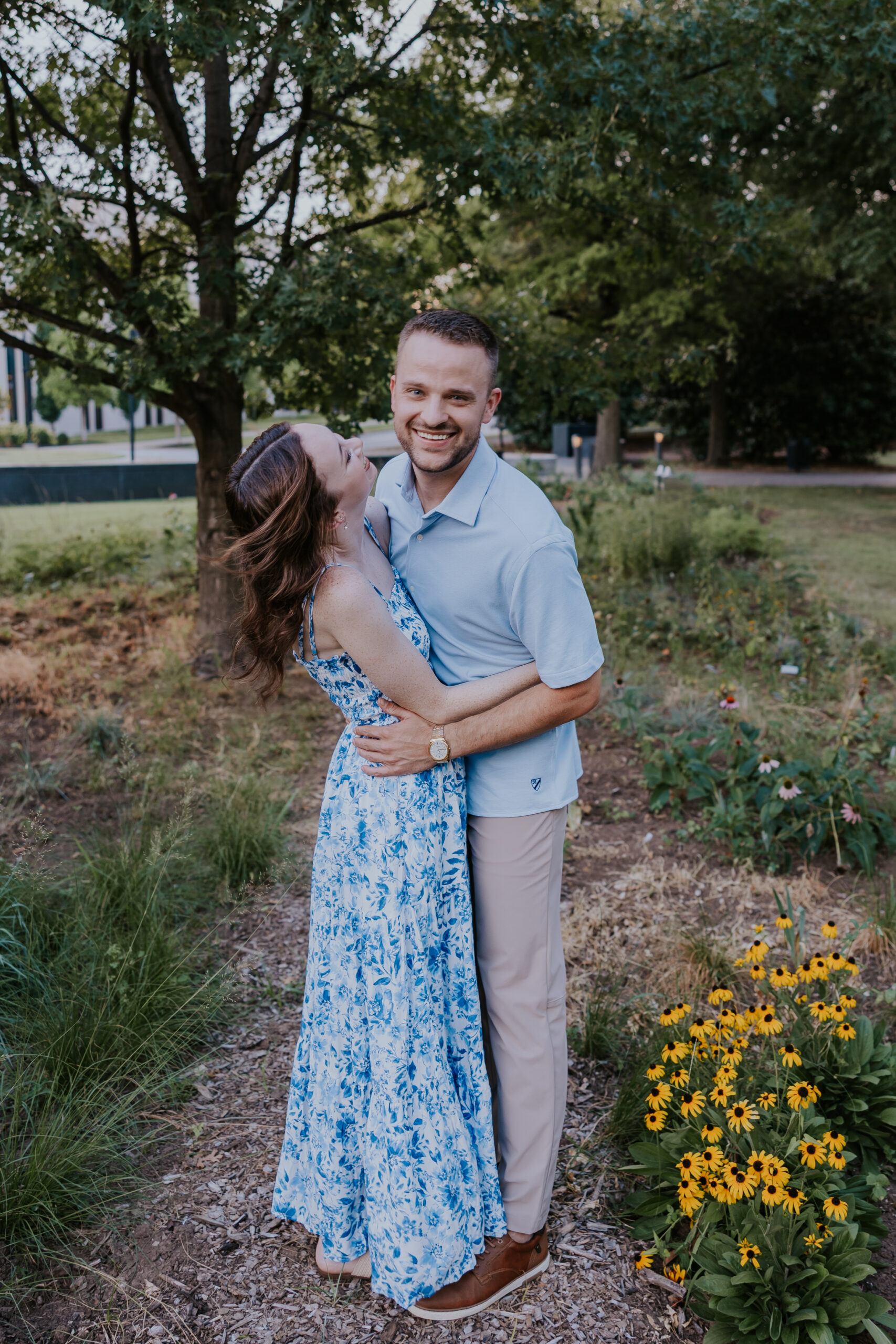 The woman hugs her smiling partner beside a bed of yellow wildflowers, both wearing comfortable and coordinated outfits.