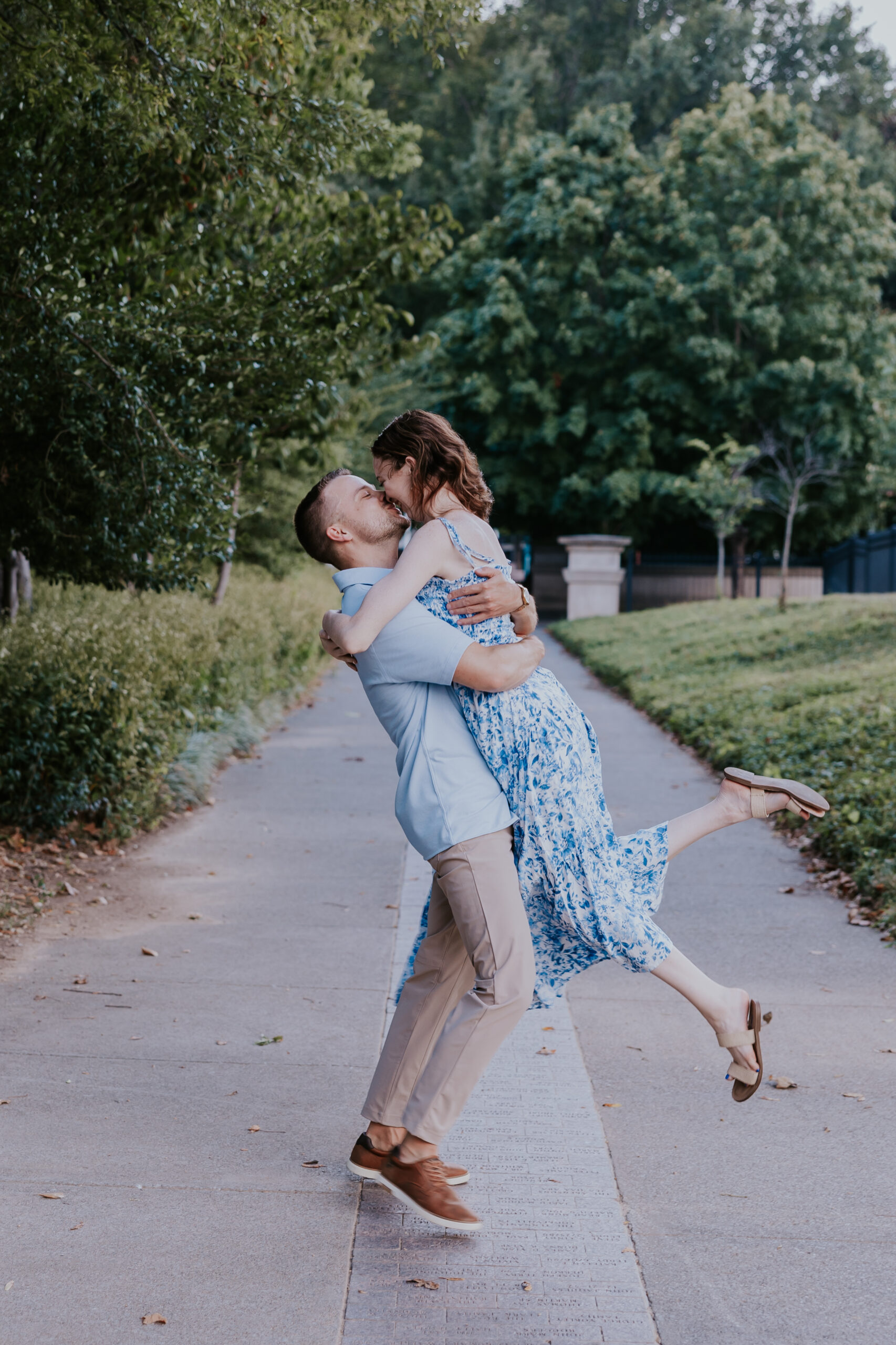 A playful lift-and-kiss moment on a shaded park path, with the couple dressed in soft blue tones perfect for engagement photos.
