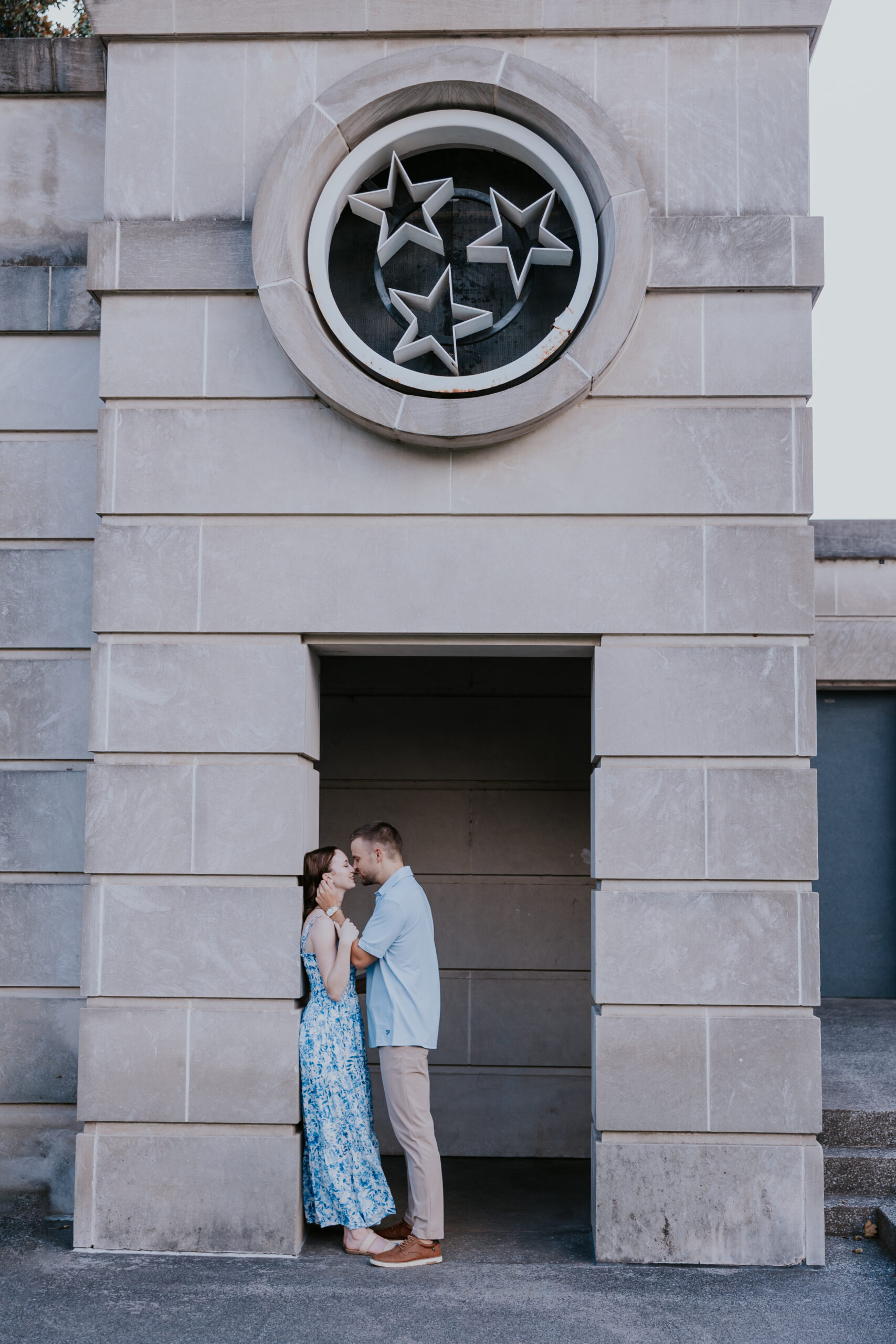 A couple embraces beneath a stone structure adorned with three stars, styled in outfits that are ideal for what to wear for engagement photos.