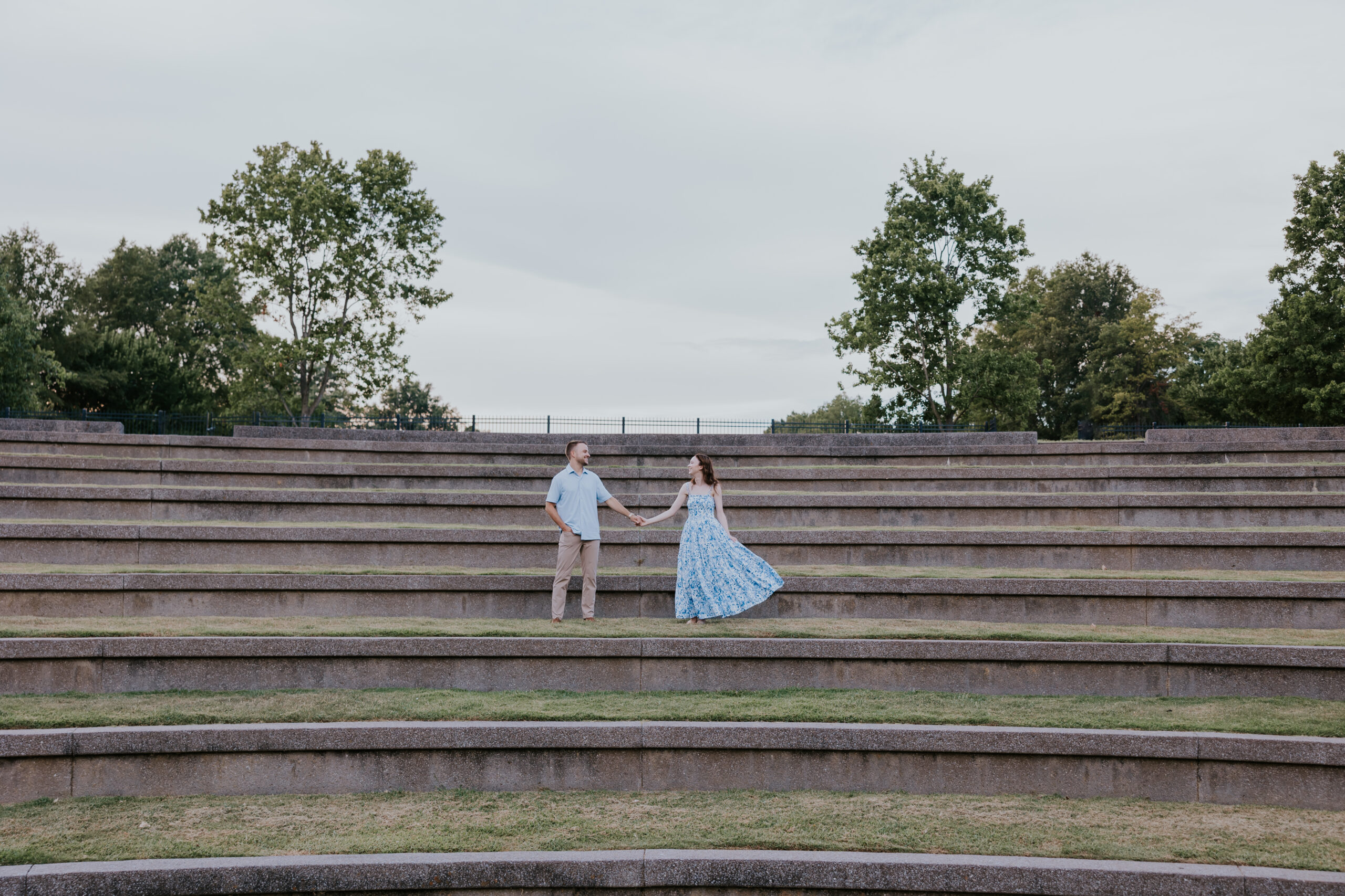 A couple holding hands on the wide stone steps of an outdoor amphitheater, captured mid-spin in flowing engagement photo attire.