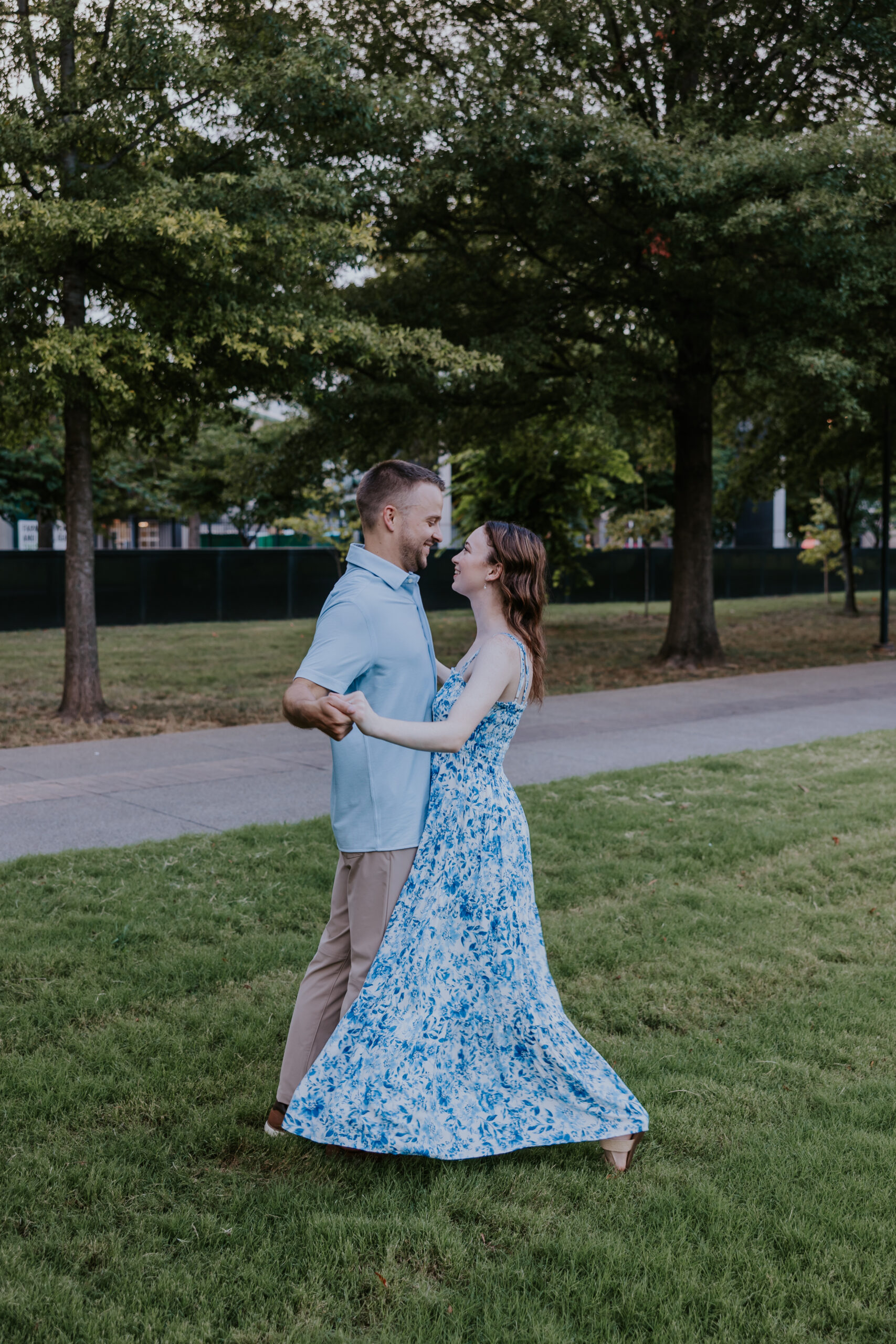 A couple dances in a park, the woman in a flowing blue floral dress and the man in a light blue shirt and khaki pants — a great example of what to wear for engagement photos.